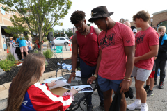 People gathered around a table at an outdoor event, signing autographs on items displayed.