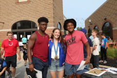Three people posing for a picture at an event. A woman in a red shirt stands between two men, one wearing a blue tank top