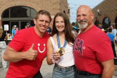 Three men posing together at an event, one holding a medal around his neck, another wearing a red shirt and giving thumbs