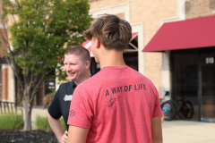 Man in red shirt standing next to another man, both smiling, wearing a hat, in front of a restaurant.