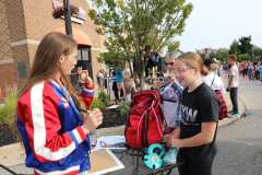 This is a color photograph featuring an outdoor scene where a woman in a red jacket and blue jeans stands at a table with a