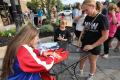 Two women at a table, one signing autographs, other looking on; both are smiling and appear to be enjoying themselves.