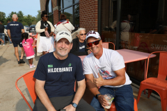 Two men posing at an outdoor event, one wearing a hat and the other a baseball cap, both smiling and standing near a table