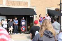 People gathered outside a building for an event, with one individual standing on a stage holding a microphone.