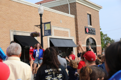 People gathered outside a restaurant, some waving, others standing in line; a woman on a stage behind the crowd, holding