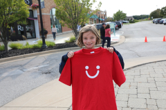 Girl holding a red t-shirt with a white smiley face on it. She is standing outside in front of a building.