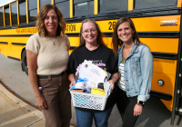 Three people standing in front of a school bus. The person on the left is holding a basket of items.