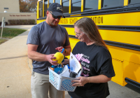 A man and woman standing next to a yellow school bus, holding a basket of items.