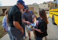 Group of people gathered outside a school building, receiving awards from a man in a blue shirt and cap.