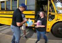 Two people standing outside a yellow school bus, one handing something to the other.