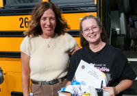 Two people standing in front of a school bus, one holding a sign that reads "241030 Bus Driver of the Month".