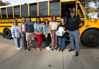 Group of people posing in front of a school bus, celebrating the driver's achievement as 'bus driver of the month'.