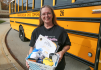 Bus driver posing with a basket of stuffed animals in front of school bus.